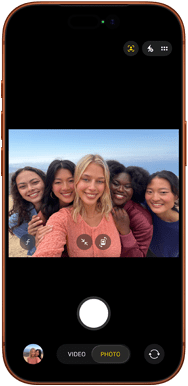 Przód iPhone’a 17 Pro w kolorze kosmicznego pomarańczu. Na ekranie kadr z aparatu przedniego Center Stage z wycentrowanym grupowym selfie pięciu osób