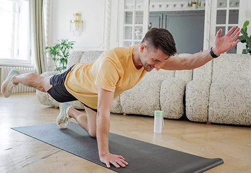 Mężczyzna w żółtej koszulce wykonuje ćwiczenie na macie w salonie. W tle sofa i roślina.