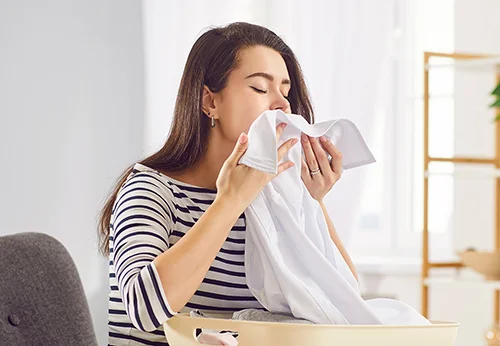 Osoba w paskowanej bluzce wącha biały materiał nad koszem na pranie w jasnym pomieszczeniu.