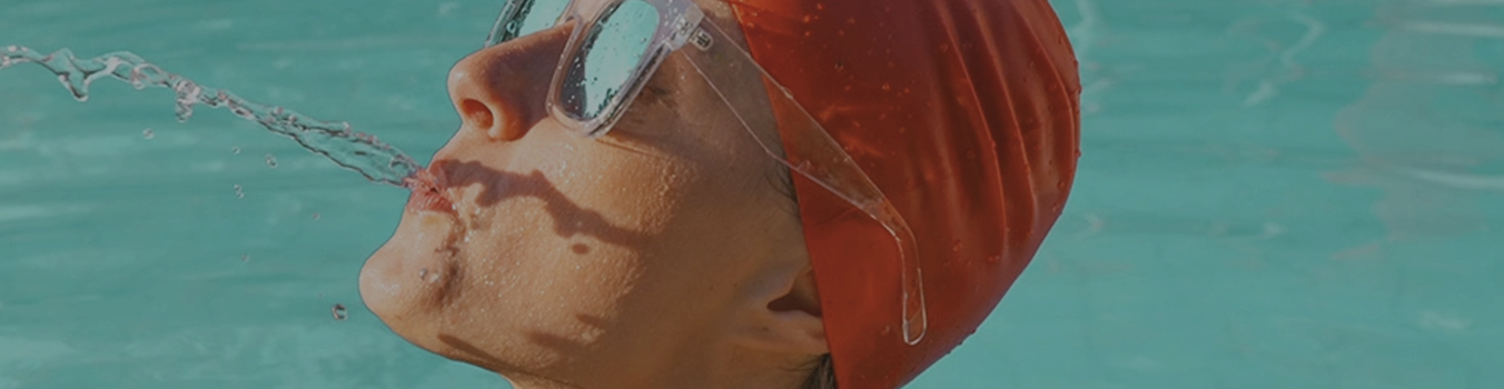 Kobieca głowa w czepku i z okularami przeciwsłonecznymi w tle z basenem wypełnionym wodą.