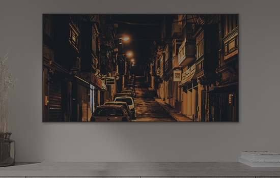 Duży telewizor wiszący w jasnym pomieszczeniu, na którego ekranie widać ciemną, wąską uliczkę miejską.