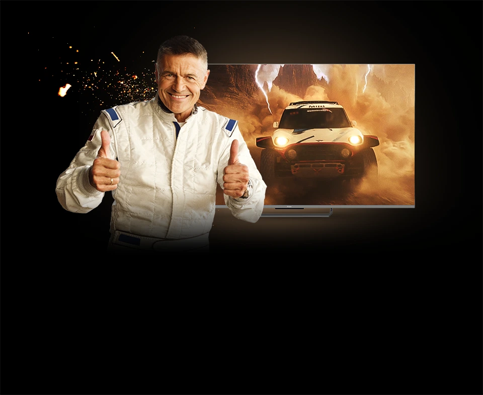 Uśmiechnięty mężczyzna w jasnym kombinezonie pokazujący uniesione kciuki na tle telewizora Haier z dynamiczną sceną samochodu terenowego