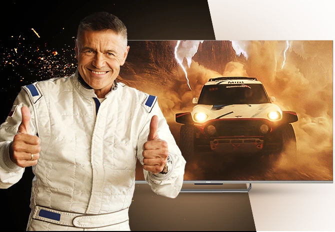 Uśmiechnięty mężczyzna w jasnym kombinezonie pokazujący uniesione kciuki na tle telewizora Haier z dynamiczną sceną samochodu terenowego