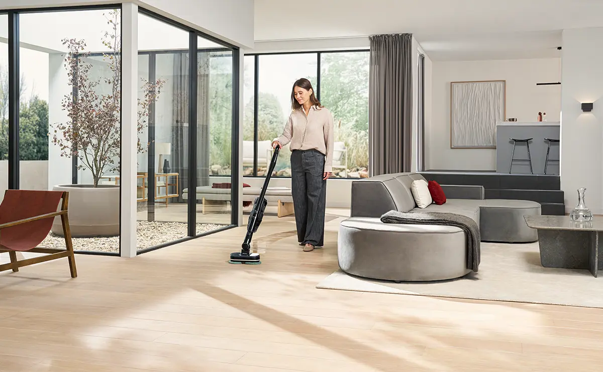 Kobieta odkurza drewnianą podłogę w jasnym salonie bezprzewodowym odkurzaczem Miele Triflex HX3