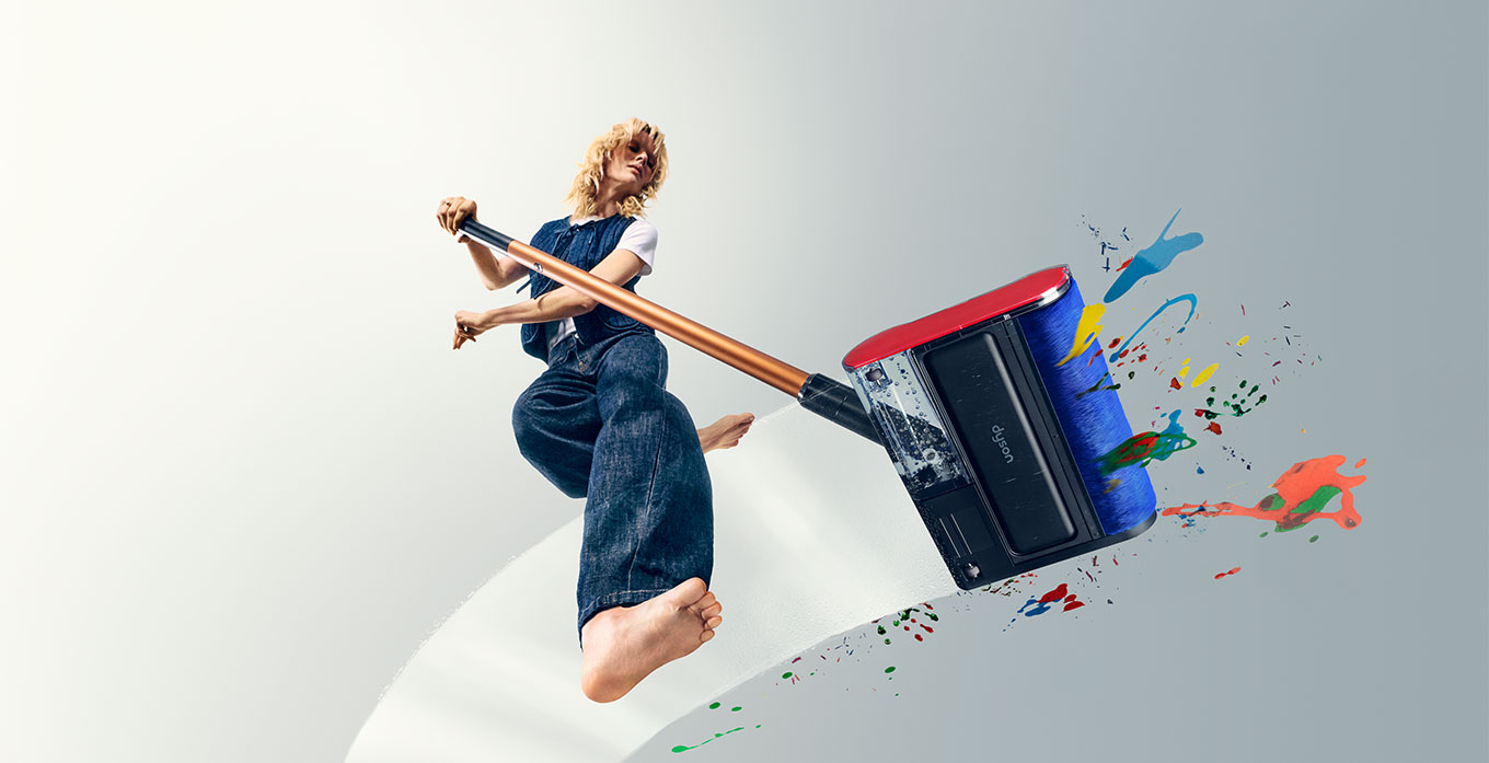 Modelka, w ujęciu od dołu, trzyma w ręku mop Dyson PencilWash i zmywa różnokolorowe plamy na przezroczystej podłodze.