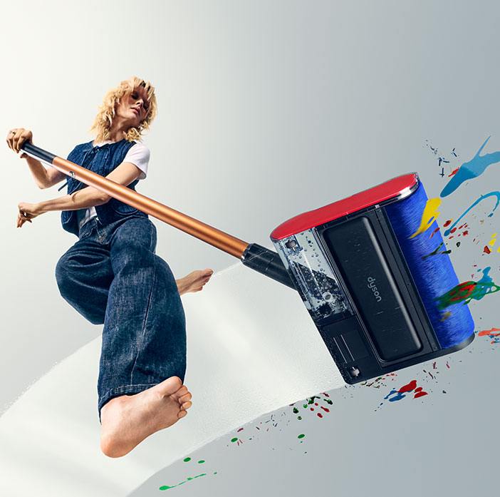 Modelka, w ujęciu od dołu, trzyma w ręku mop Dyson PencilWash i zmywa różnokolorowe plamy na przezroczystej podłodze.
