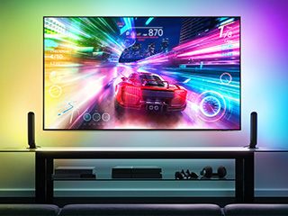 Telewizor wyświetlający dynamiczną grę wyścigową, otoczony kolorowym podświetleniem LED na ścianie oraz dwoma smukłymi głośnikami po bokach.