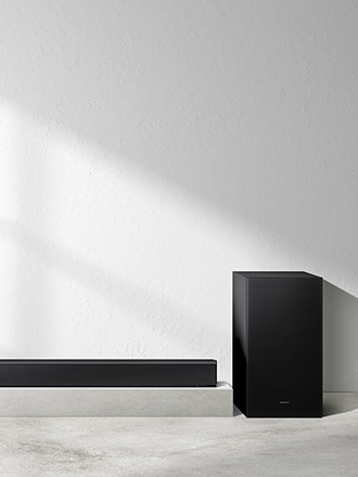 Czarny subwoofer stojący obok fragmentu soundbara Samsung HW-B650F na tle minimalistycznej, jasnej ściany z efektem światłocienia.