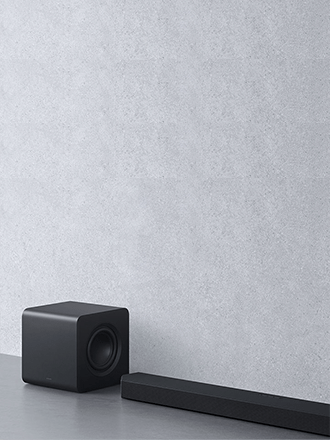 Czarny, sześcianowy subwoofer ustawiony tuż za podłużną listwą soundbara Samsung HW-Q800F na tle jasnoszarej, strukturalnej ściany.
