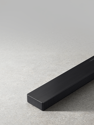 Smukły, podłużny czarny soundbar Samsung HW-QS700F z metalową kratką, sfotografowany z bliska pod kątem na szarym, betonowym podłożzu.