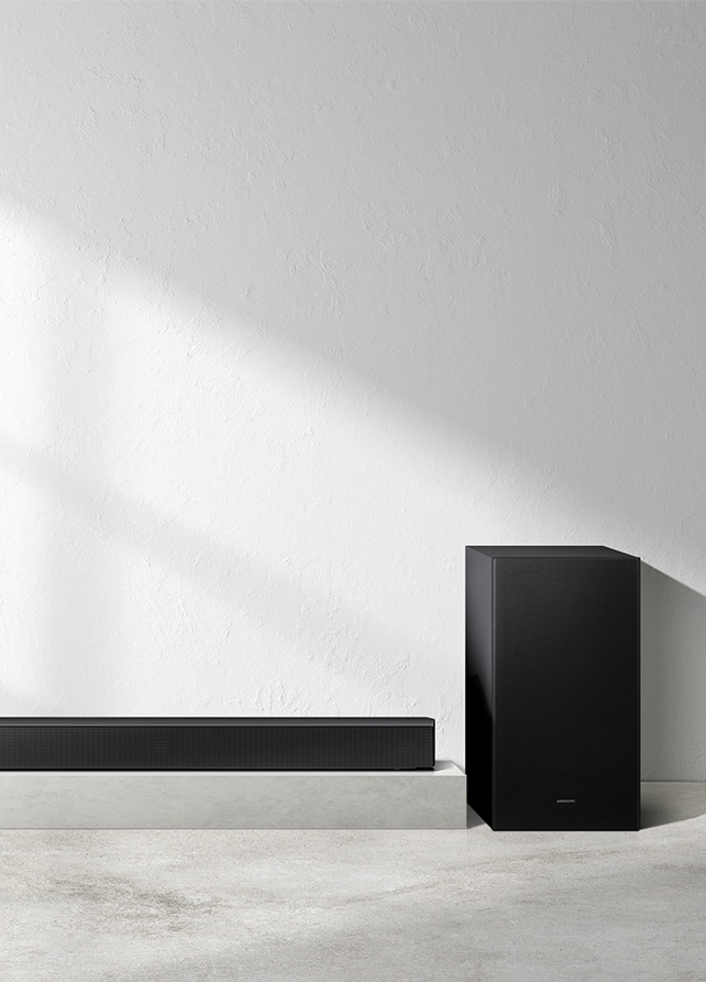 Czarny subwoofer stojący obok fragmentu soundbara Samsung HW-B650F na tle minimalistycznej, jasnej ściany z efektem światłocienia.