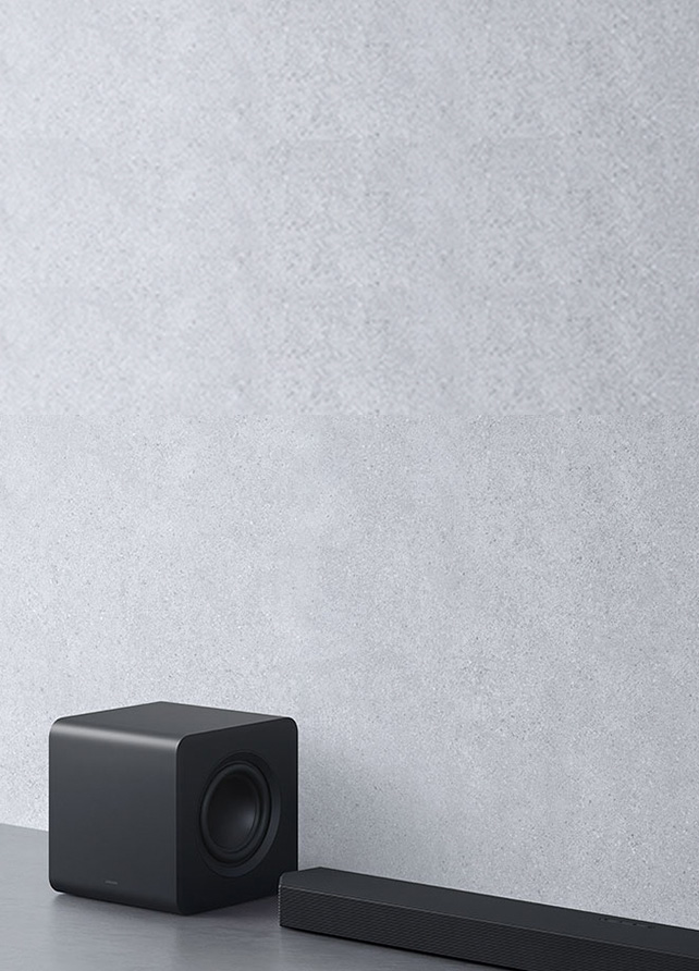 Czarny, sześcianowy subwoofer ustawiony tuż za podłużną listwą soundbara Samsung HW-Q800F na tle jasnoszarej, strukturalnej ściany.