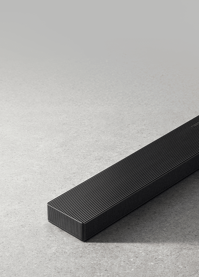 Smukły, podłużny czarny soundbar Samsung HW-QS700F z metalową kratką, sfotografowany z bliska pod kątem na szarym, betonowym podłożzu.