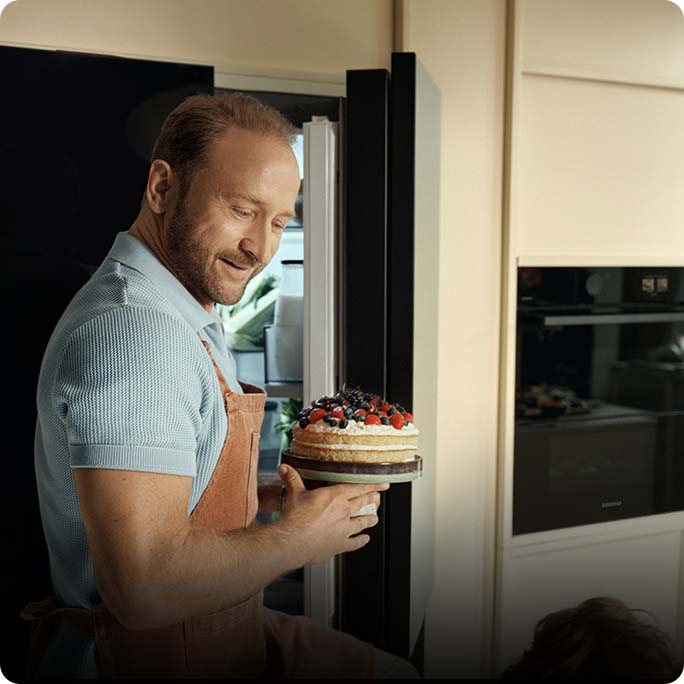 U&sacute;miechni&eogon;ty Borys Szyc w fartuchu kuchennym wyjmuje z lod&oacute;wki ma&lstrok;y tort z owocami.
