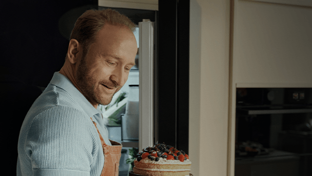 U&sacute;miechni&eogon;ty Borys Szyc w fartuchu kuchennym wyjmuje z lod&oacute;wki ma&lstrok;y tort z owocami.