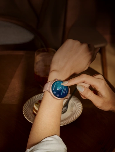 Zegarek HUAWEI Watch 6 Active różowy na kobiecym nadgarstku, kobieta palcem klika w niebieski ekran, poniżej talerz z ciastem